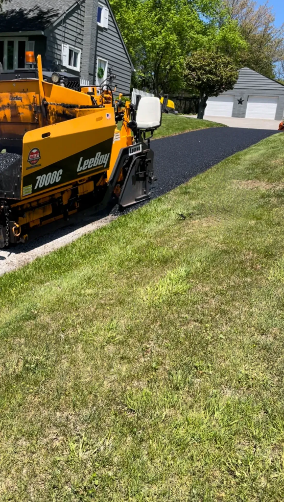 A yellow LeeBoy 7000C paving machine is laying fresh asphalt on a residential driveway next to a grassy lawn.