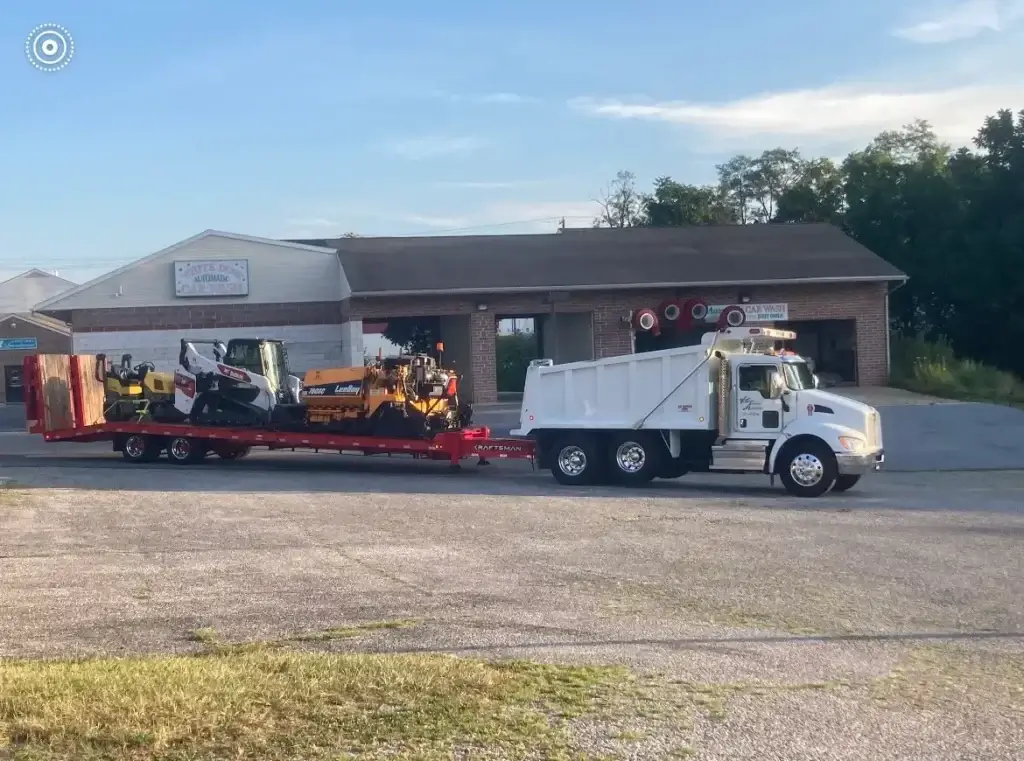 A white dump truck is parked in front of a building, towing a red trailer loaded with construction equipment.