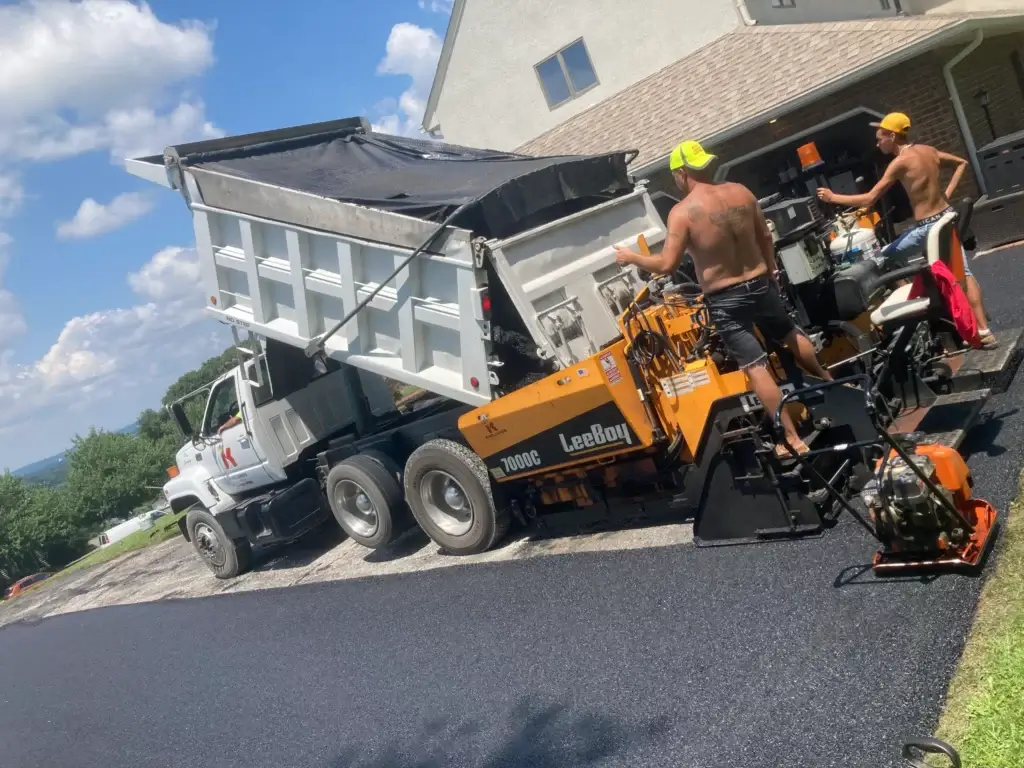 Two workers operate paving equipment as asphalt is poured from a dump truck onto a driveway outside a house on a sunny day.