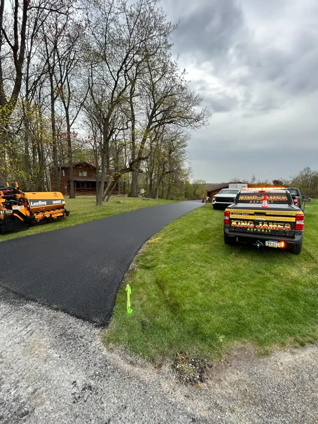 Freshly paved asphalt driveway with paving equipment and a work truck parked on the grass beside it; trees and a house are visible in the background under a cloudy sky.