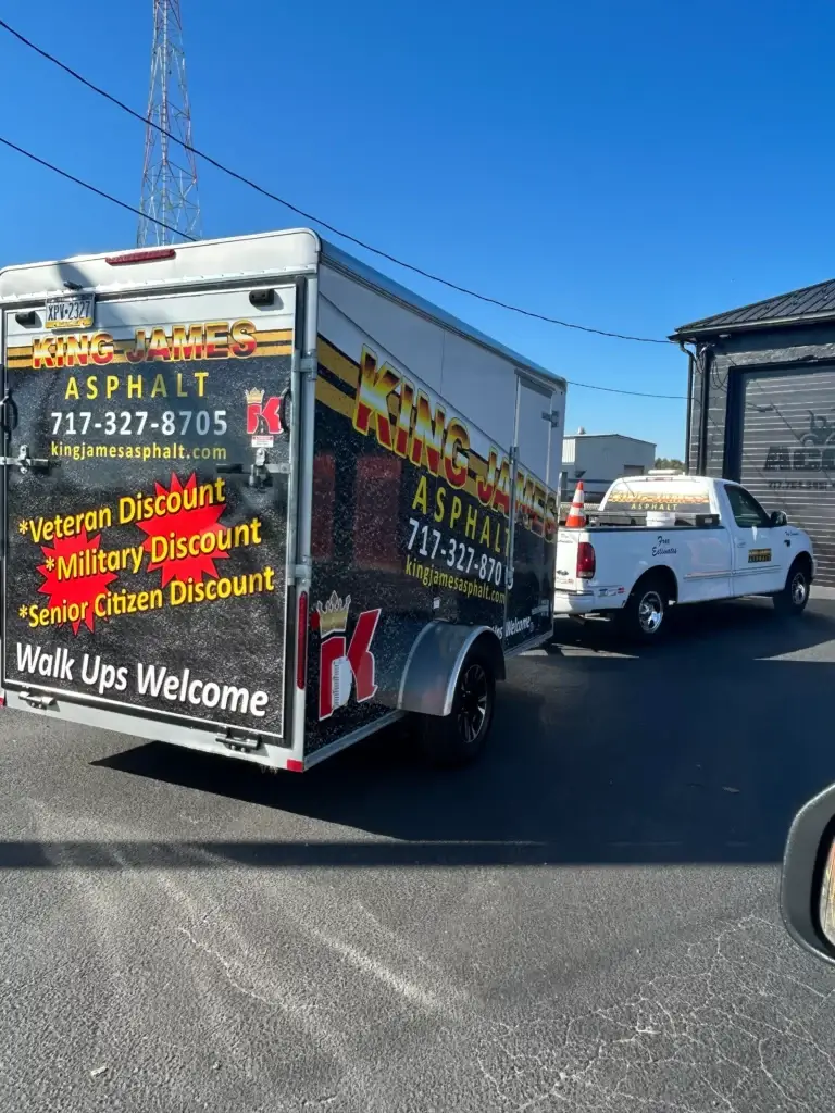 Truck and trailer for King James Asphalt parked on asphalt, displaying contact info and discounts for veterans, military, and seniors. Another work truck is visible in the background.