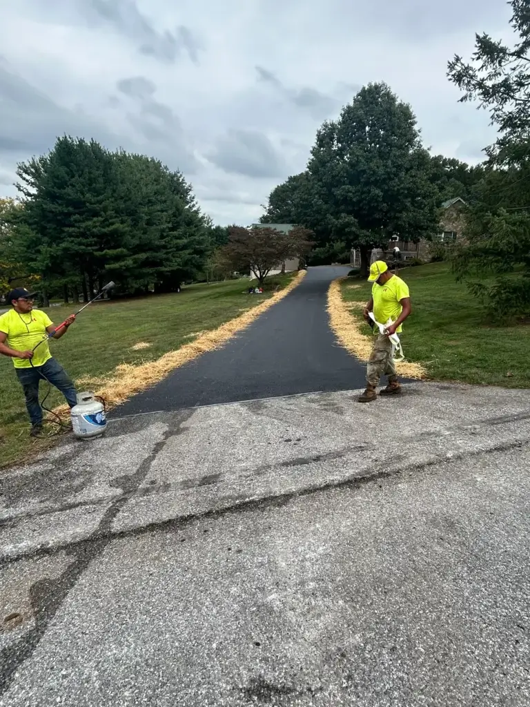 Two workers in yellow shirts stand next to a freshly paved driveway, applying straw along the edges; trees and houses are visible in the background.
