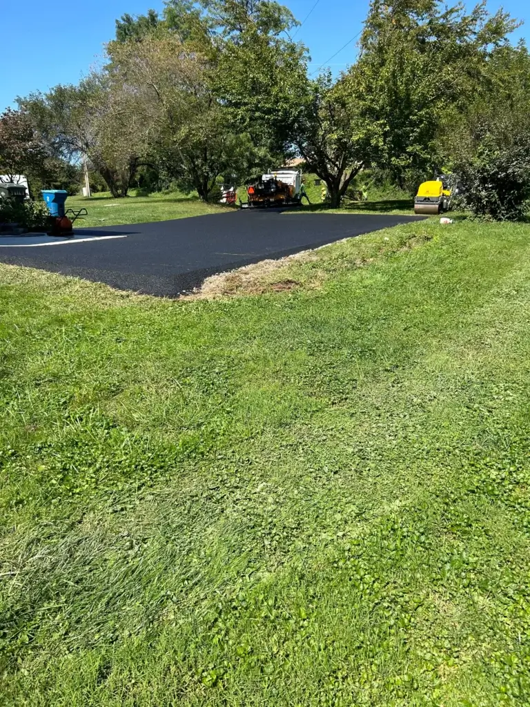 Workers are paving a section of asphalt surrounded by green grass and trees under a clear blue sky. Equipment and vehicles are visible near the work area.