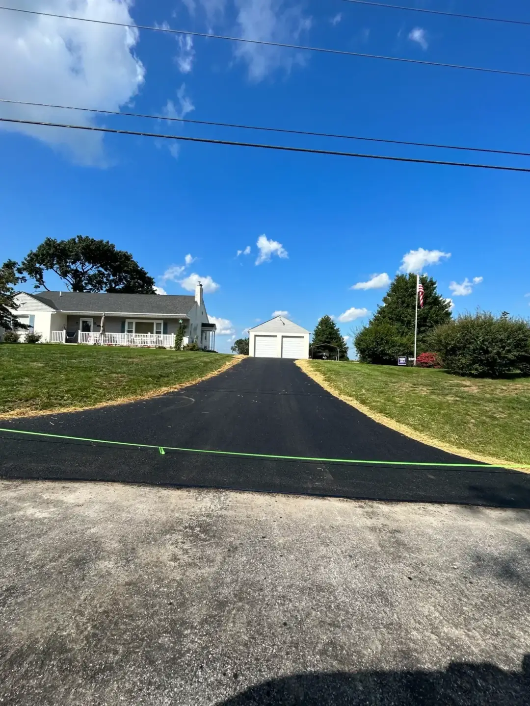 A newly paved asphalt driveway leads up to a white house with a detached garage, surrounded by grass and trees under a blue sky with scattered clouds.