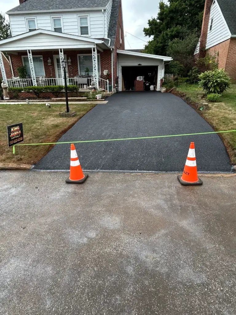 Freshly paved driveway of a house, blocked off by orange traffic cones and a green cord, with a sign indicating wet asphalt.