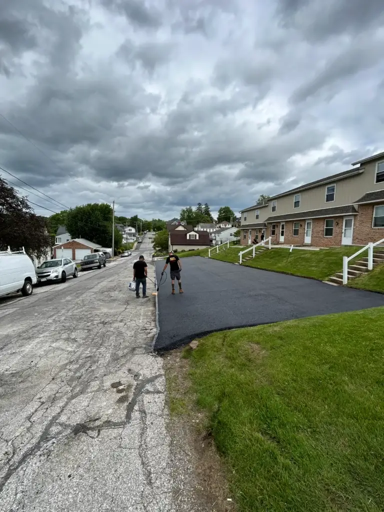 Two people stand on a newly paved driveway next to a residential street under a cloudy sky, with houses and parked vehicles visible.